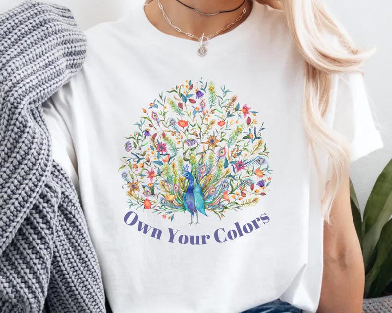 May include: White t-shirt featuring a colorful peacock design with the text "Own Your Colors". The peacock is surrounded by flowers and leaves in shades of blue, green, yellow, and purple. The shirt is a casual wear item.