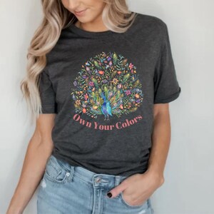 May include: Dark gray t-shirt featuring a colorful peacock design with the text "Own Your Colors" in pink. The peacock has blue, green, and yellow feathers, surrounded by floral elements. The shirt is made of a soft material.