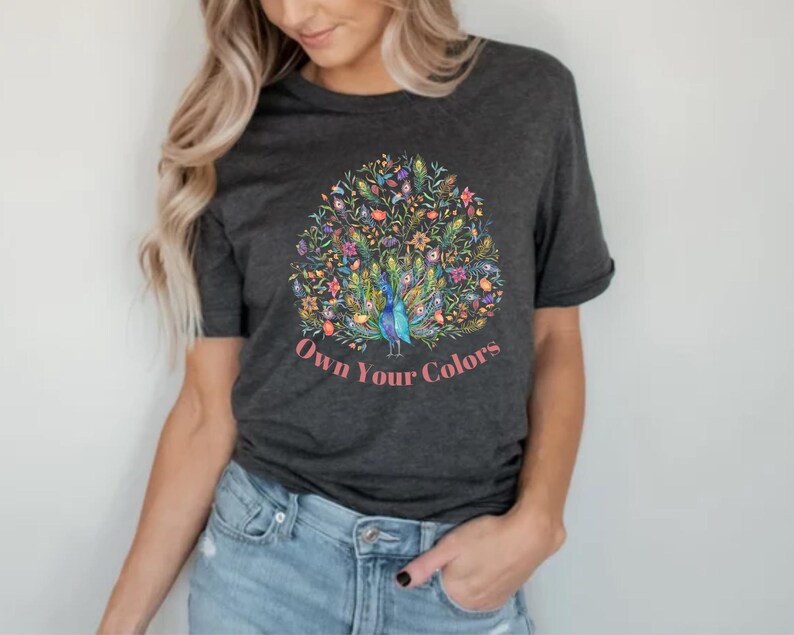 May include: Dark gray t-shirt featuring a colorful peacock design with the text "Own Your Colors" in pink. The peacock has blue, green, and yellow feathers, surrounded by floral elements. The shirt is made of a soft material.