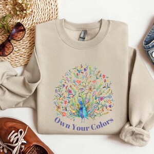 May include: Beige crewneck sweatshirt with a colorful peacock design and the text "Own Your Colors". The peacock is surrounded by flowers and leaves in shades of blue, green, pink, and yellow. The sweatshirt is made of a soft material.