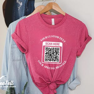May include: Heather raspberry-colored t-shirt with rolled sleeves. The shirt features a white QR code graphic with the text "YOUR CUSTOM TEXT SCAN HERE YOUR SPECIAL MESSAGE". The shirt is hanging on a wooden hanger.