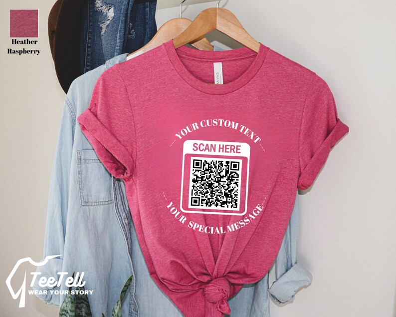 May include: Heather raspberry-colored t-shirt with rolled sleeves. The shirt features a white QR code graphic with the text "YOUR CUSTOM TEXT SCAN HERE YOUR SPECIAL MESSAGE". The shirt is hanging on a wooden hanger.