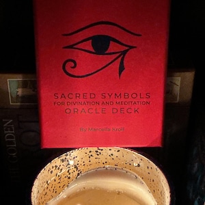 May include: A red box with the words "SACRED SYMBOLS FOR DIVINATION AND MEDITATION ORACLE DECK" and an eye symbol. Below the box is a lit candle in a speckled container.