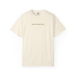 Könnte beinhalten: Cremefarbenes Kurzarm-T-Shirt mit dem Text "MATTHÄUS 13:8" in Braun aufgedruckt. Das Shirt hat einen Rundhalsausschnitt und ein Etikett am Nacken. Das T-Shirt ist aus einem weichen Material gefertigt.