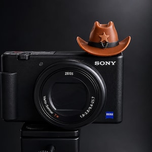 以下が含まれることがあります： 赤い三脚に取り付けられた、Zeiss Vario-Sonnar T* 1.8-2.8/9.4-25.7レンズ付きの黒いソニー製デジタルカメラ。黒いバンドと星のエンブレムが付いた小さな茶色のカウボーイハットが、レンズの上のカメラ本体の天面に置かれています。