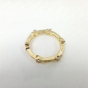 Puede incluir: Un anillo dorado con un dise&ntilde;o geom&eacute;trico repetitivo. El anillo presenta peque&ntilde;as piedras preciosas transparentes y est&aacute; sobre un fondo blanco. La palabra "Elegance" es visible en el anillo.