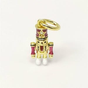 May include: A gold-colored charm in the shape of a nutcracker soldier. The charm has a red and gold uniform with a star on the hat. The charm has a small loop at the top for attaching to a bracelet or necklace. The soldier's feet are white.