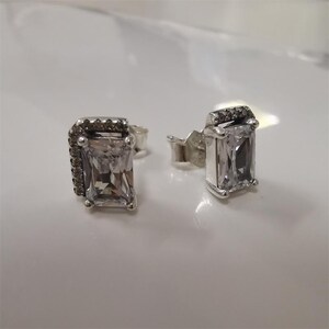 Peut inclure: Une paire de boucles d'oreilles à clous en argent avec des pierres précieuses rectangulaires, claires et à facettes. Une boucle d'oreille a une rangée de petites pierres sombres sur le bord supérieur. Les boucles d'oreilles sont présentées sur une surface blanche.