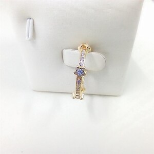 Puede incluir: Un anillo dorado con una gema azul en forma de estrella y peque&ntilde;as gemas transparentes. El anillo se muestra sobre una superficie blanca. El dise&ntilde;o presenta una estrella en el centro y piedras m&aacute;s peque&ntilde;as a lo largo de la banda.