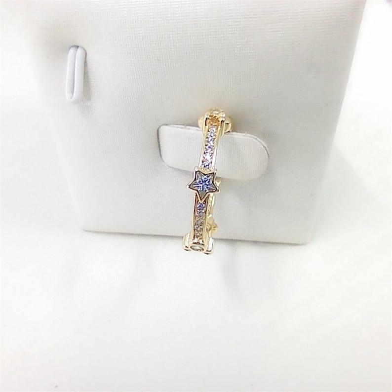 Puede incluir: Un anillo dorado con una gema azul en forma de estrella y peque&ntilde;as gemas transparentes. El anillo se muestra sobre una superficie blanca. El dise&ntilde;o presenta una estrella en el centro y piedras m&aacute;s peque&ntilde;as a lo largo de la banda.