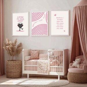 Pu&ograve; includere: Una scena di asilo nido con tre stampe incorniciate con disegni rosa e bianchi e citazioni ispiratrici. Sono presenti anche una culla bianca con biancheria da letto rosa e una sedia a baldacchino rosa. Un cesto di vimini contiene una pianta.