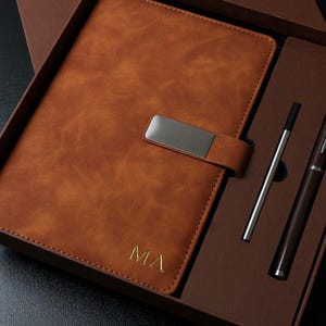 May include: A brown leather journal and pen set in a gift box. The journal has a silver clasp and the initials "MA" in gold. The set includes a silver pen and a dark brown pen. The box is brown.
