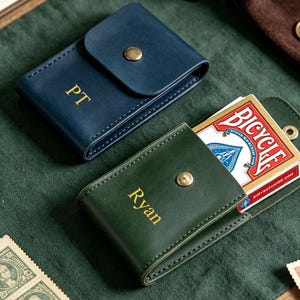 May include: Two leather card cases with gold-tone snap closures. One case is dark blue with the gold embossed letters PT, and the other is dark green with the gold embossed name Ryan, containing a deck of Bicycle playing cards inside.