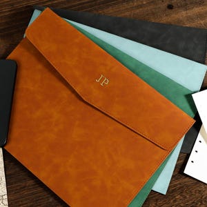 May include: A selection of leather document holders in shades of orange, green, blue, and black. The orange holder is embossed with the initials "JP" in gold. A black smartphone and map fragments are also present.