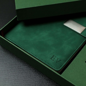 Puede incluir: Un juego de regalo verde que incluye un diario de cuero, un broche plateado y un bolígrafo a juego, presentado en una caja verde. El diario está grabado con las iniciales "EV".