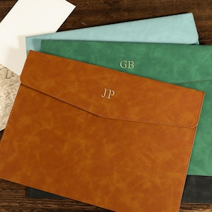 Può includere: Portadocumenti in pelle marrone, verde e blu. Il portadocumenti marrone è impresso con le iniziali "JP", e quello verde ha "GB". Buste sono visibili sullo sfondo. Su una superficie di legno.