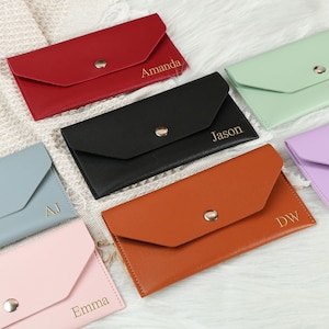 May include: A collection of personalized leather wallets in various colors, including red, black, and brown. Each wallet features a snap closure and is embossed with a name or initials in gold lettering. The wallets are arranged on a soft, white surface.