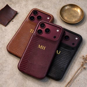 May include: Three smartphone cases with a textured lizard-skin pattern in brown, burgundy, and black. Each case features a camera cutout with a dark red frame and gold-coloured monogrammed initials. The cases are arranged in a diagonal overlapping row.