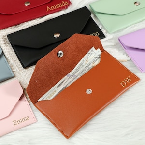 May include: An open, brown leather wallet with a snap closure, containing several $100 bills. The wallet is personalized with the initials "DW" in gold. Other colored wallets, including red, black, pink, and green, are visible in the background, each with a name.
