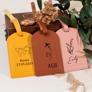 May include: Three leather luggage tags in yellow, brown, and pink. The yellow tag has a world map graphic and the name "Monika" with a date. The brown tag features an airplane and the initials "AGB". The pink tag has a floral design and the name "Emily".