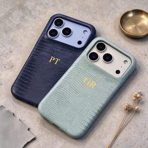 May include: Two smartphone cases with a textured lizard-skin pattern. One case is dark navy blue with the gold-embossed initials PT, and the other is light sage green with the gold-embossed initials GR. Both feature cutouts for triple camera lenses.