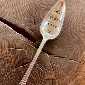 May include: A silver spoon with the words "Your quote here" stamped on the bowl, and a small heart symbol. The spoon is set against a textured, dark brown wooden surface.