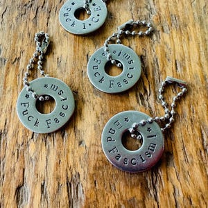 Puede incluir: Cuatro colgantes de metal plateado con cadenas de bolas. Cada colgante es un c&iacute;rculo plano con un agujero en el centro y texto estampado. El texto incluye las palabras "Fuck Fascism" y "End Fascism".