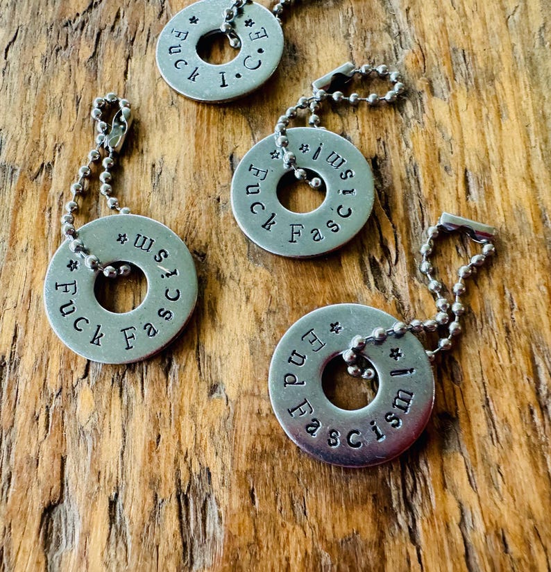 Puede incluir: Cuatro colgantes de metal plateado con cadenas de bolas. Cada colgante es un c&iacute;rculo plano con un agujero en el centro y texto estampado. El texto incluye las palabras "Fuck Fascism" y "End Fascism".