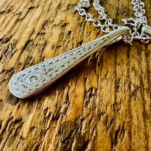 May include: Silver pendant necklace featuring an engraved spoon design. The pendant has an elongated shape with intricate patterns and a circular motif at the base. The silver chain sits on a textured wooden surface.