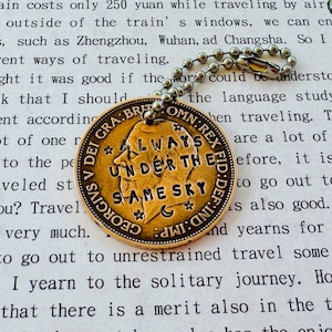 May include: A gold-colored coin pendant with the text "ALWAYS UNDER THE SAME SKY" and a portrait of a man. The coin is attached to a silver ball chain. The background is a page of printed text.
