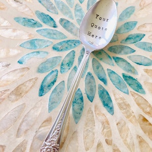 May include: A silver spoon with the text "Your Quote Here" stamped on the bowl, along with a small heart. The spoon has a floral design on the handle and rests on a mosaic surface with blue and white accents.