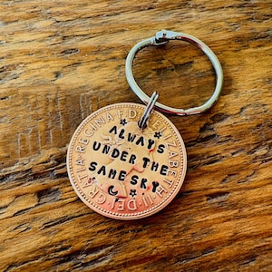 May include: A copper coin keychain with the text "Always Under The Same Sky." The coin is attached to a silver keyring. The background is a wooden surface.