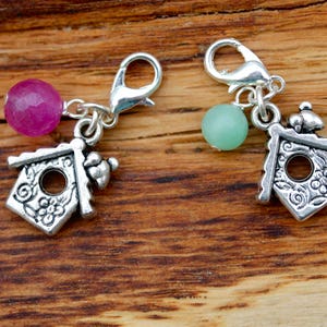 May include: Two silver charm pendants with birdhouse designs. Each pendant has a small bird on top and a gemstone bead. One bead is pink and the other is green.