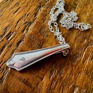 Könnte beinhalten: Eine silberne Löffel-Anhänger-Halskette mit einer filigranen Kette. Der Löffel-Anhänger hat einen dekorativen Rand und einen kleinen eingravierten Buchstaben "P". Die Halskette liegt auf einer rustikalen Holzoberfläche.