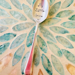 May include: A silver spoon with the words "My Little Spoon" engraved on the bowl. The spoon is on a background of teal and brown mosaic tiles.