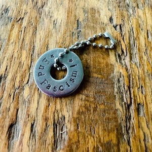Puede incluir: Un colgante circular de color plateado con las palabras "End Fascism!" estampadas alrededor del borde. El colgante est&aacute; unido a un collar de cadena de bolas. El fondo es una superficie de madera.