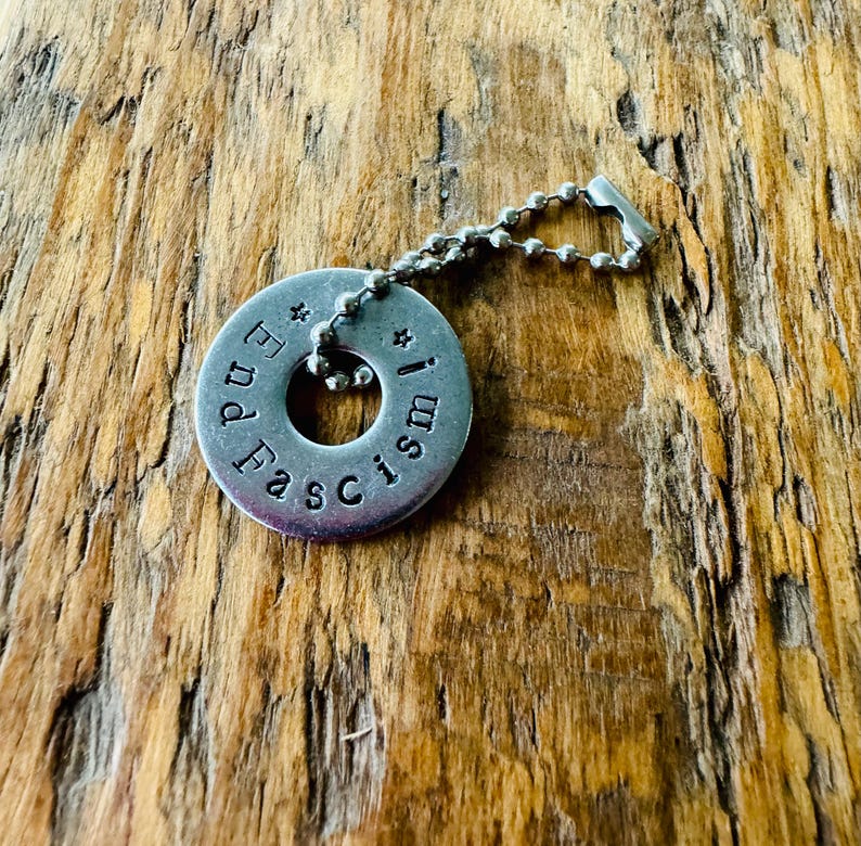 Puede incluir: Un colgante circular de color plateado con las palabras "End Fascism!" estampadas alrededor del borde. El colgante est&aacute; unido a un collar de cadena de bolas. El fondo es una superficie de madera.