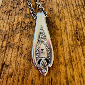 May include: Silver pendant necklace crafted from a spoon head. The pendant showcases floral detailing and the letter "E". The necklace has a silver chain and is presented on a wooden surface.