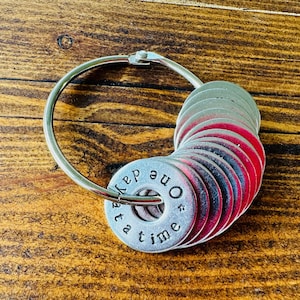 May include: A silver metal ring holds a collection of small, round, metal discs. The discs are grey with red edges, and the first disc has the words "one day at a time" stamped on it. The background is a wooden surface.