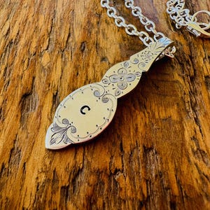 May include: Silver-toned pendant necklace with an engraved floral design and the letter "C". The pendant is attached to a silver chain. The necklace is displayed on a wooden surface.