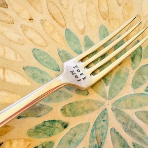 May include: A silver fork with the words "Fork Me!" engraved on the handle. The fork is lying on a decorative plate with a floral pattern.