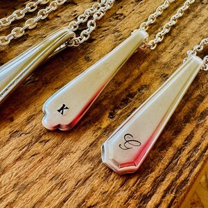 May include: Silver spoon necklaces with chain links. Two spoons are engraved with the letters "K" and "G". The spoons have a decorative edge and are set against a rustic wooden background.