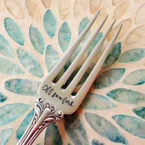 May include: A silver fork with four tines, engraved with the phrase "Off you fork" in a script font. The fork has a decorative handle and is set against a mosaic background with teal and beige leaf-like patterns.