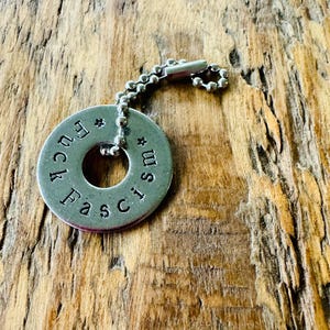 Puede incluir: Un colgante de llavero de metal plateado con las palabras "Fuck Fascism" estampadas alrededor del borde. El colgante es circular con un agujero en el centro y est&aacute; unido a una cadena de bolas.