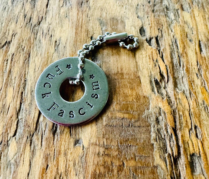 Puede incluir: Un colgante de llavero de metal plateado con las palabras "Fuck Fascism" estampadas alrededor del borde. El colgante es circular con un agujero en el centro y est&aacute; unido a una cadena de bolas.
