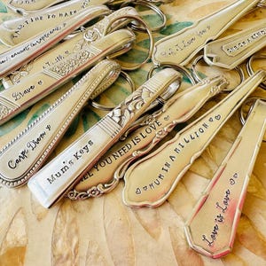 以下が含まれることがあります： 「Love is Love」や「Mum's Keys」などのフレーズが刻まれた銀のスプーンのキーホルダーのコレクション。 スプーンは装飾的な縁があり、銀色のキーリングに付いています。 背景は明るい色の表面です。