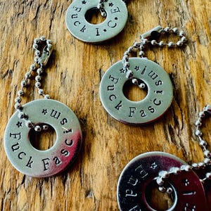Puede incluir: Primer plano de varios colgantes de metal plateado con un dise&ntilde;o circular y un agujero en el centro. Cada colgante tiene las palabras "Fuck Fascism" estampadas, junto con una cadena. Los colgantes est&aacute;n sobre una superficie de madera.