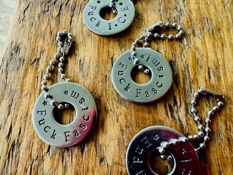Puede incluir: Primer plano de varios colgantes de metal plateado con un dise&ntilde;o circular y un agujero en el centro. Cada colgante tiene las palabras "Fuck Fascism" estampadas, junto con una cadena. Los colgantes est&aacute;n sobre una superficie de madera.