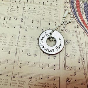 May include: A silver pendant with the text "Your Wording Here" around a central hole, suspended from a beaded chain. The pendant is set against a backdrop of vintage sheet music, with a partial view of a hot air balloon.