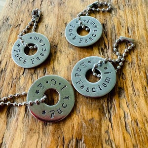 Puede incluir: Cuatro colgantes plateados con cadenas de bolas. Cada colgante es un c&iacute;rculo plano con un agujero en el centro y texto estampado que dice "Fuck Fascism" o "End Fascism!". Los colgantes se muestran sobre una superficie de madera.